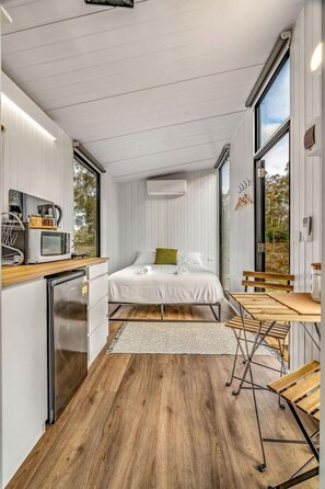 A modern bedroom with a bed, bedside table, and a view of the outdoors. - Cozy Tiny House with Barbecue, Firepit and River Views in Victoria, Australia (Dadswells Bridge)