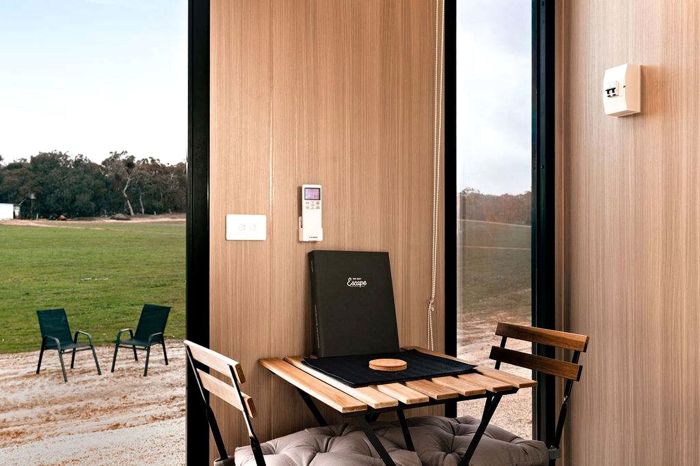 A room with a wooden table, two chairs, a black chair, and a view of a grassy field through a window.