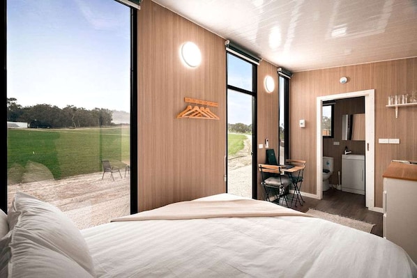 A modern hotel room with a large bed, a dining area with a table and chairs, and a view of a green landscape.
