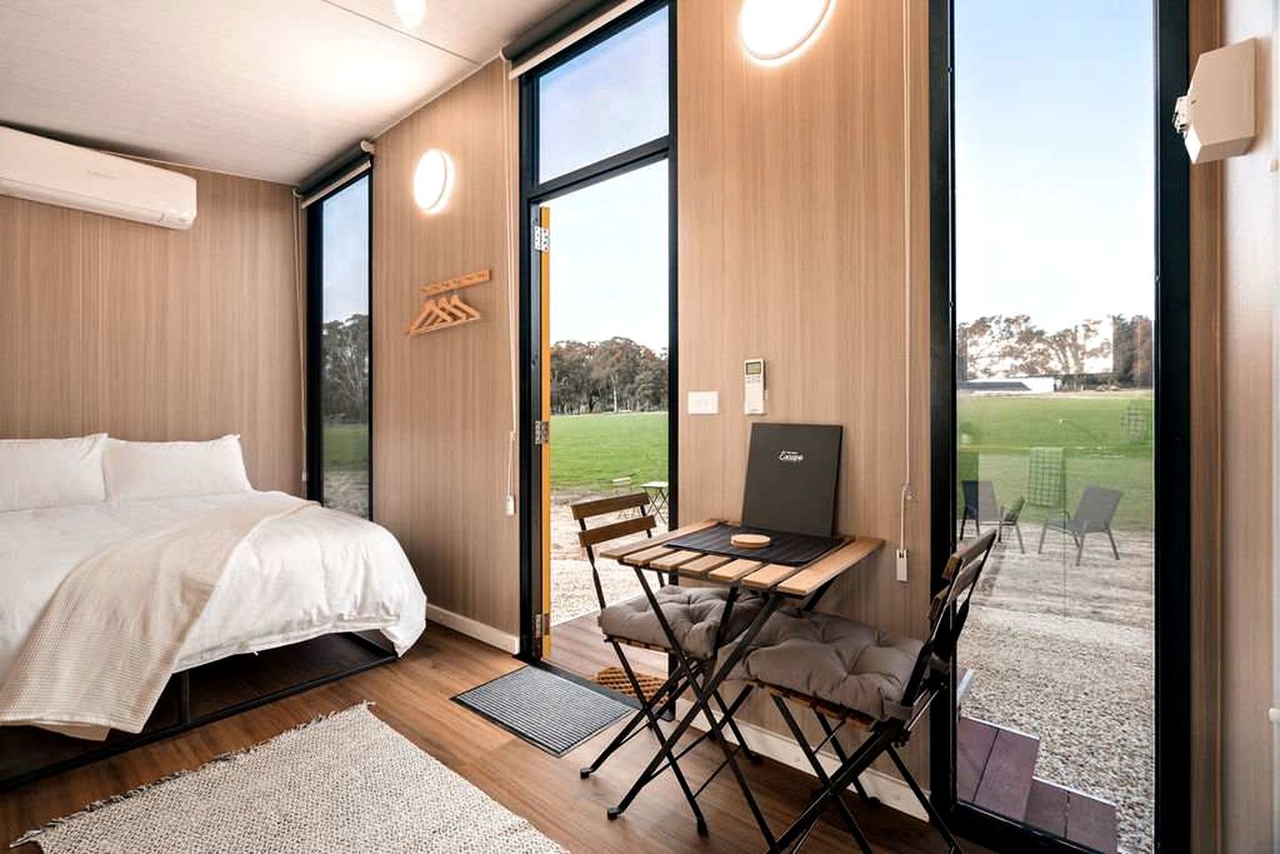 A modern bedroom with a bed, a desk, and a laptop, featuring large windows and a view of a grassy area.