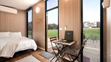A modern bedroom with a bed, a desk, and a laptop, featuring large windows and a view of a grassy area.
