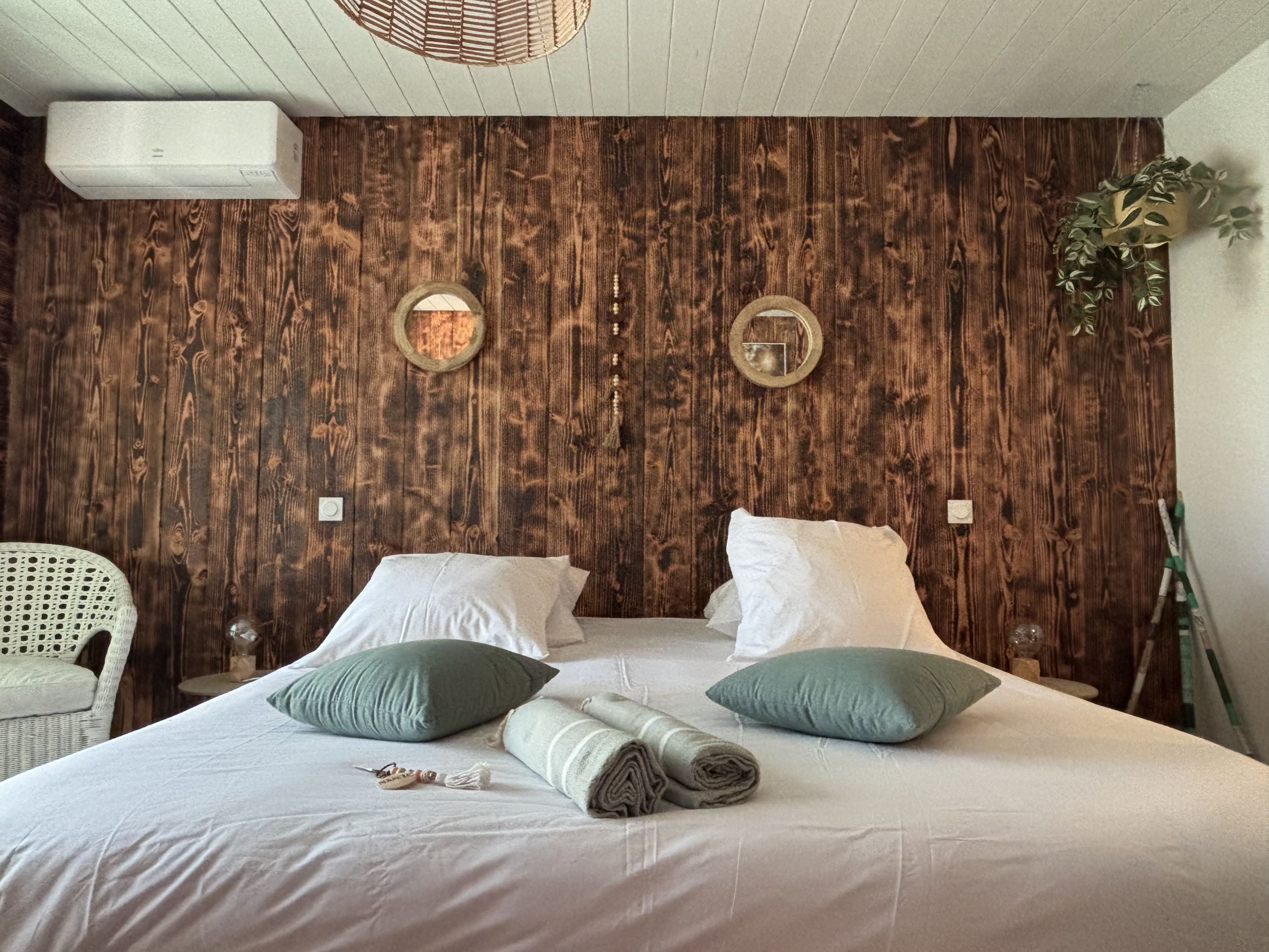 Un dormitorio con pared de madera, cama con almohadas blancas y verdes, una silla blanca y una planta.