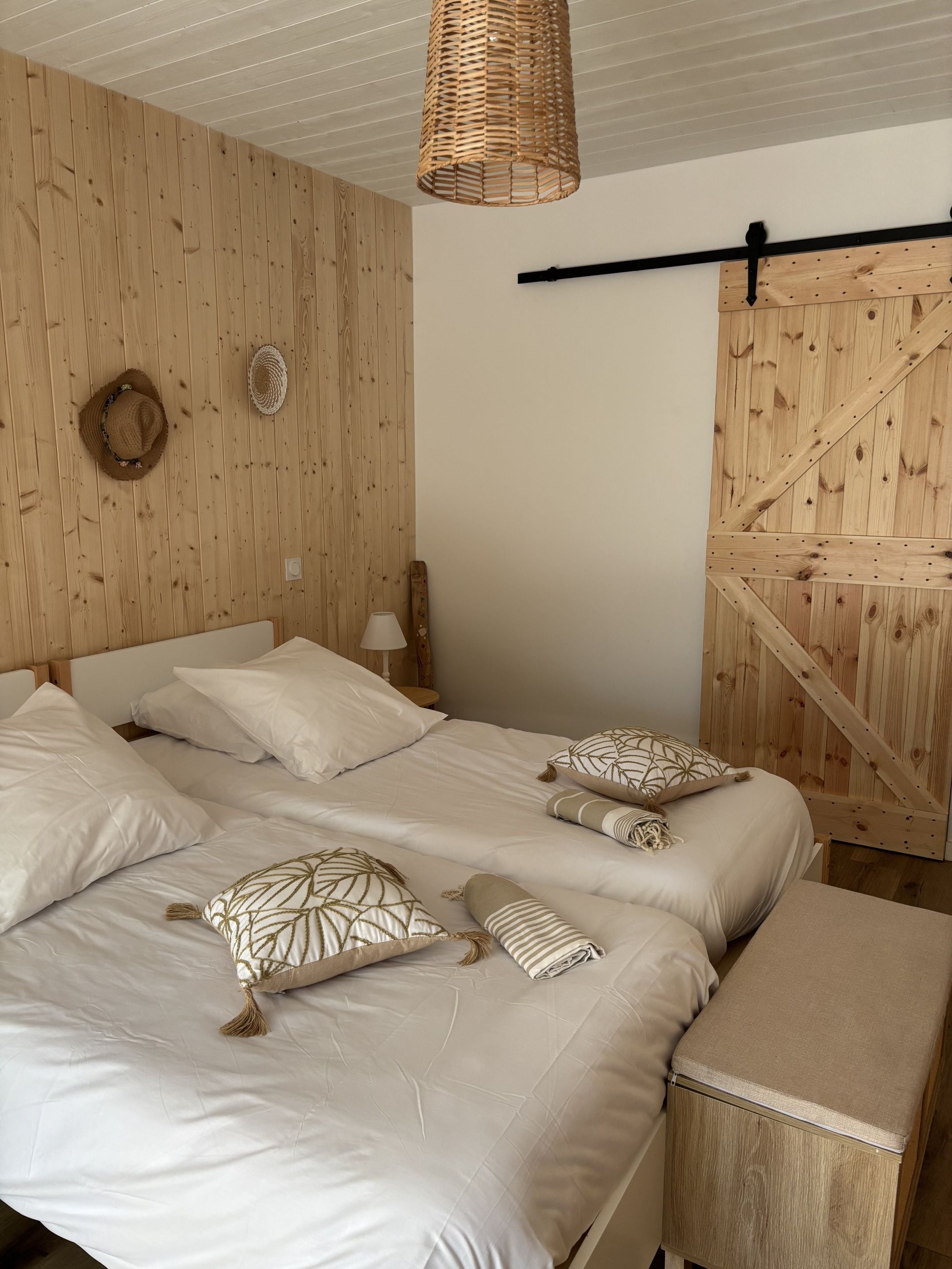 Un dormitorio con pared de listones de madera, cama con ropa de cama blanca, lámpara de techo trenzada y un banco de madera.