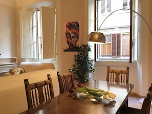 A dining area with a table, chairs, a vase with flowers, a painting on the wall, and a window with shutters. - Battistero (Florence)