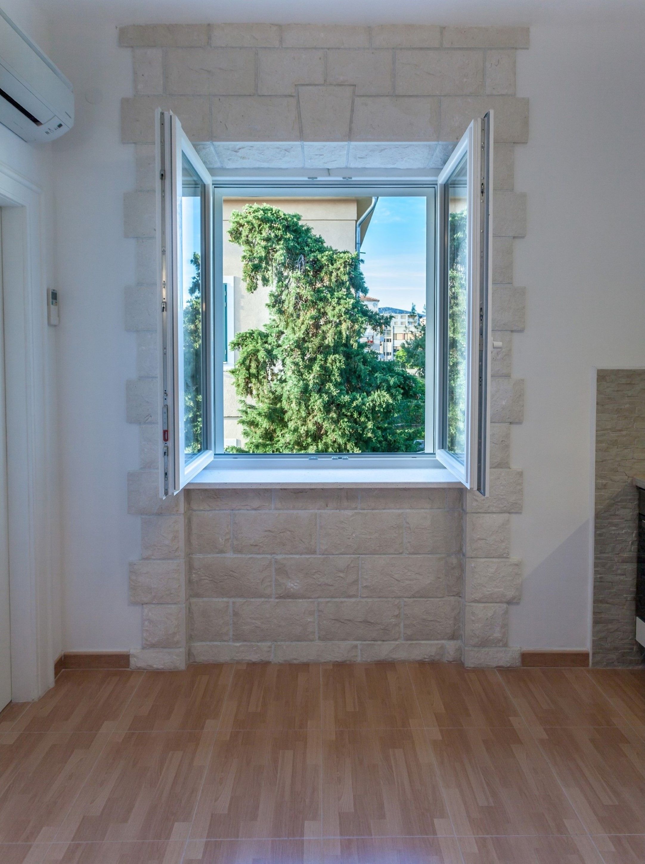 Een kamer met een groot raam, omlijst door stenen blokken, een houten vloer en uitzicht op groen en gebouwen.