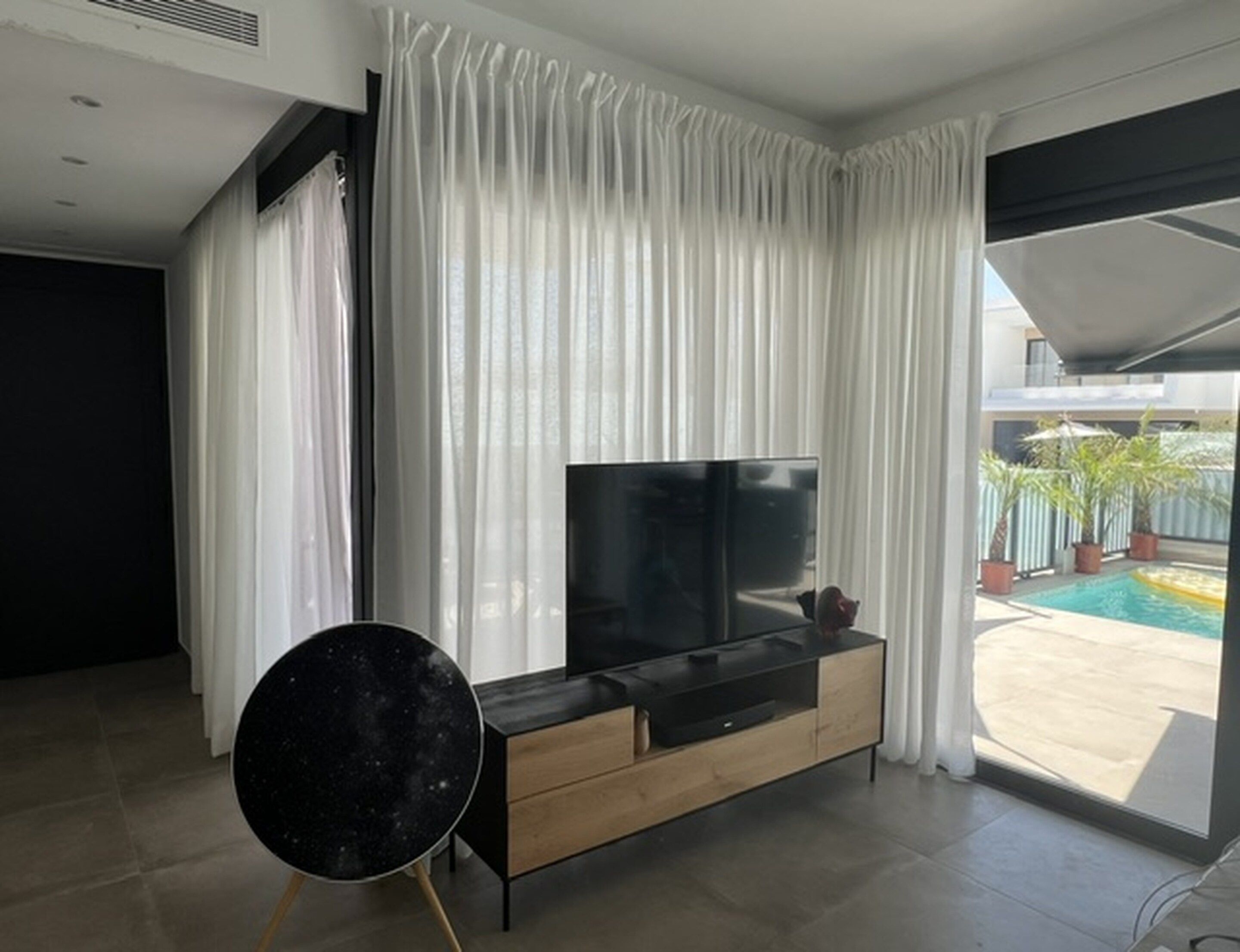 A modern living room with a flat-screen TV, a black round table, and a sliding glass door leading to a pool area.
