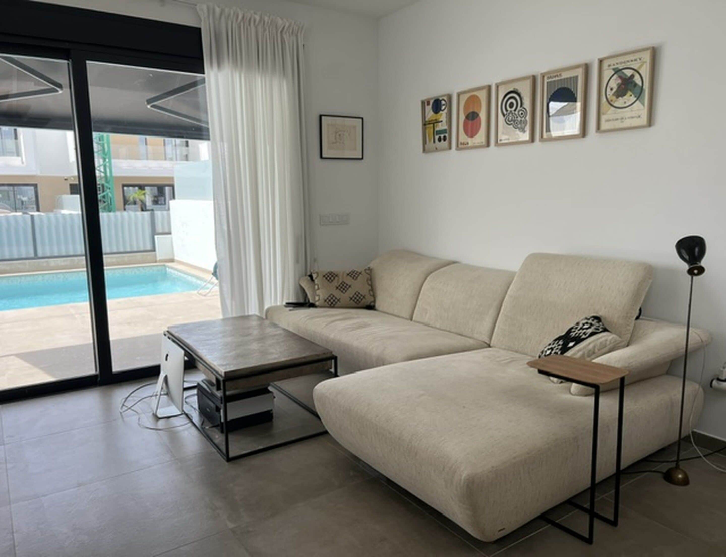 A modern living room with a beige sofa, a coffee table, and a floor lamp. There is a pool visible through the glass door.