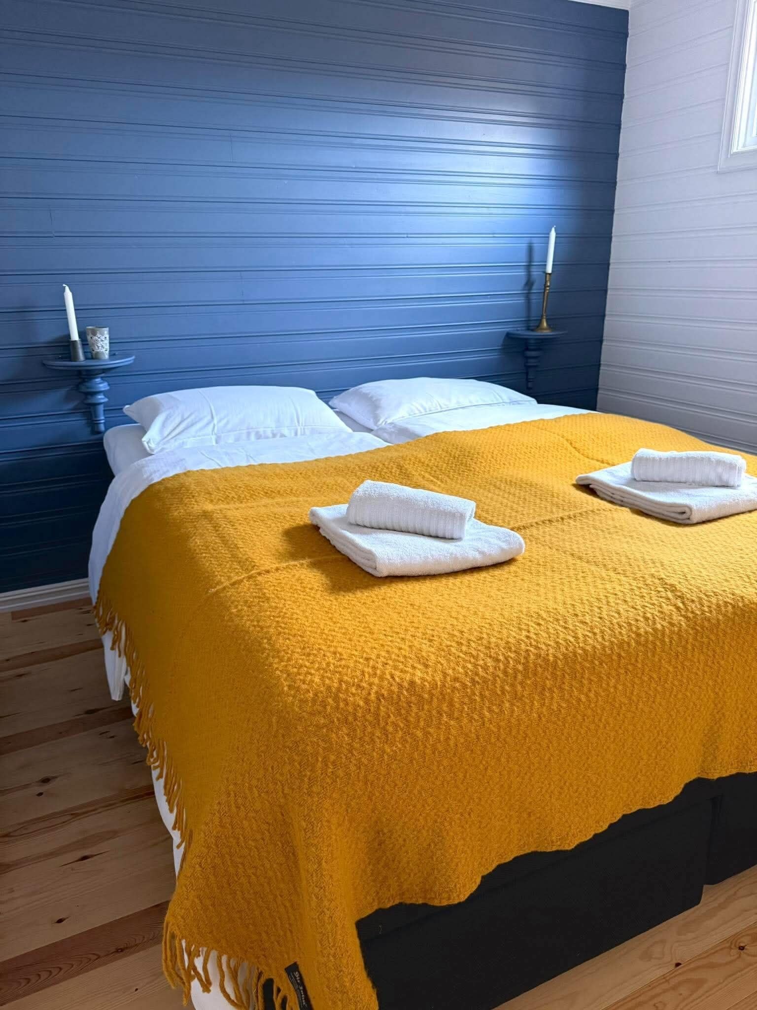 Een bed met een gele deken en twee witte handdoeken, een blauw hoofdeinde en twee nachtkastjes met kaarsen.