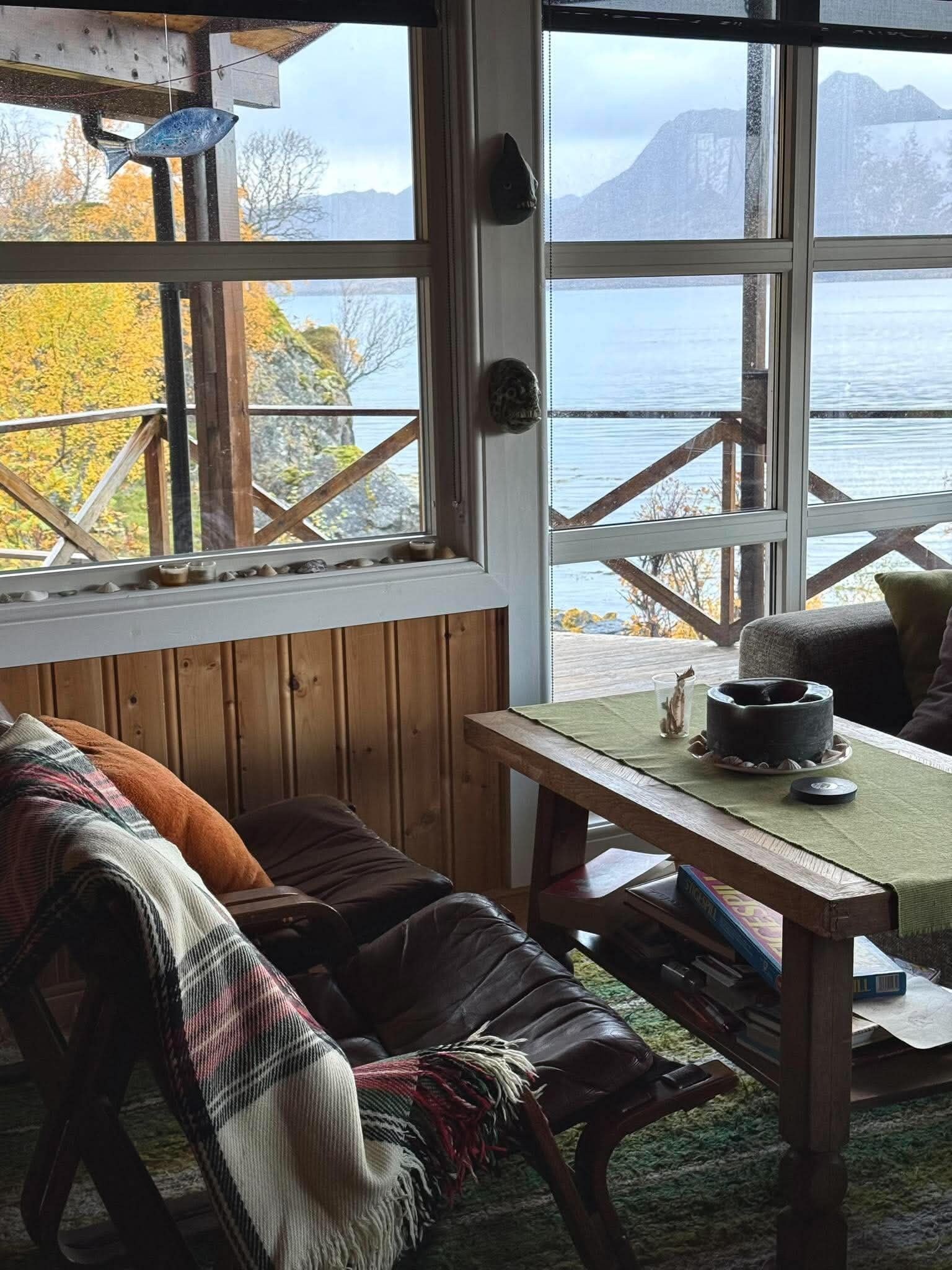 A cozy room with a wooden bench, a table with a cup and a book, and a window overlooking a body of water and mountains.