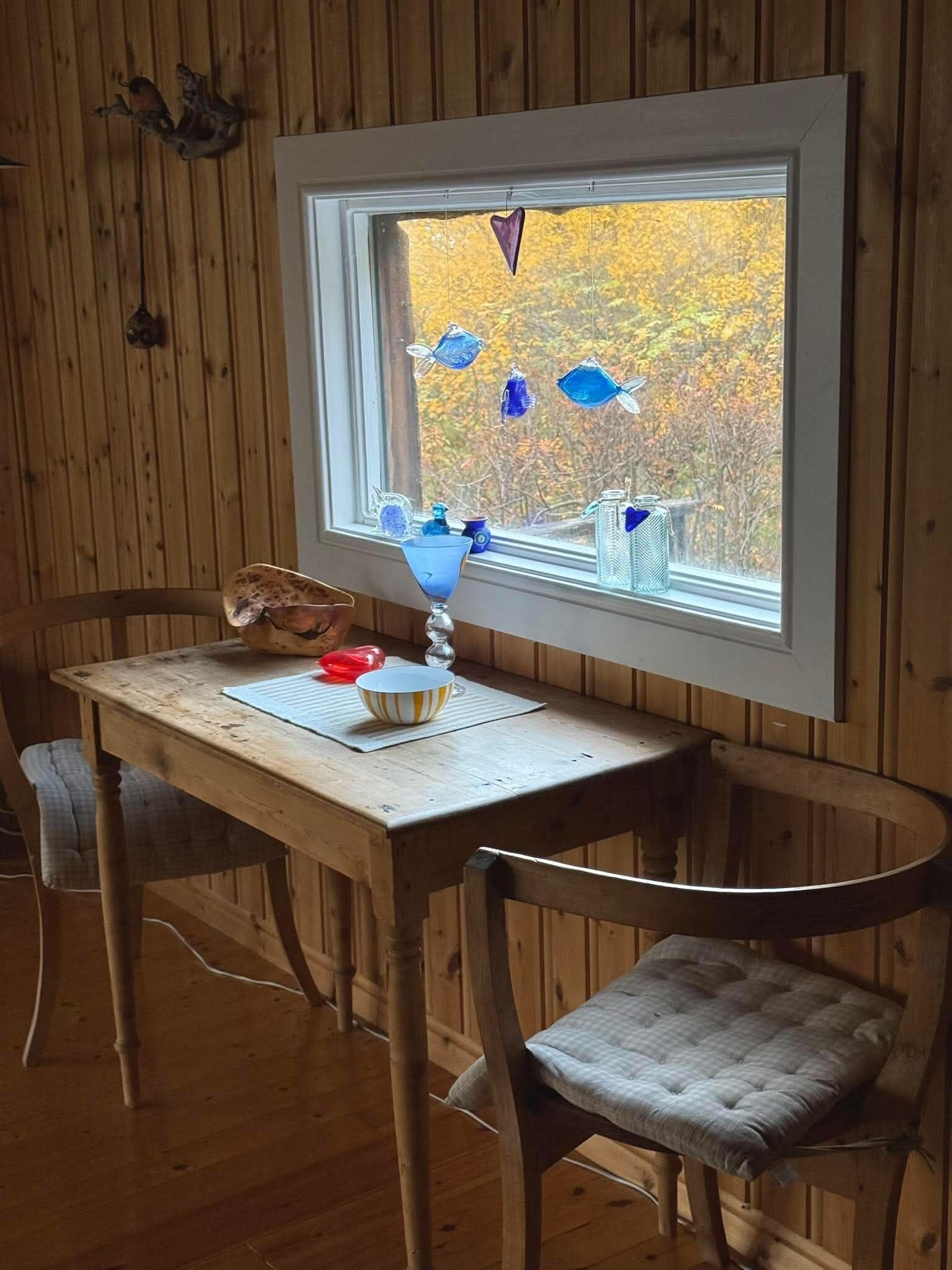 Una mesa de madera con un cuenco y una lámpara, dos sillas y una ventana decorada con adornos de peces de vidrio azul.