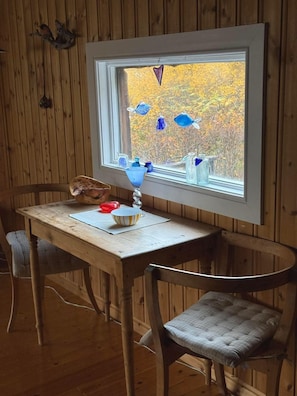 Una mesa de madera con un cuenco y una lámpara, dos sillas y una ventana decorada con adornos de peces de vidrio azul.