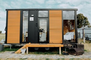 A modern mobile home with a wooden exterior, a visible kitchen area, and a small outdoor seating space.
