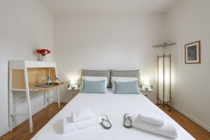 Una habitació d'hotel amb un llit, una taula petita amb una ampolla i un got, un vas amb flors i un parell de xanclas al llit.