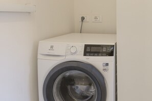 Eine Waschmaschine mit Frontlader, einer Bedienkonsole, einer Steckdose und einem an der Wand befestigten Regal.