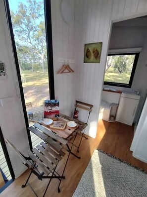 A room with a wooden floor, a window, a hanging coat rack, a small table with a box and a cup, and a bench.