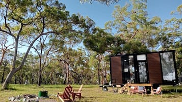 Une cabane moderne et minimaliste, dotée d’une grande fenêtre et entourée d’un paysage naturel composé d’arbres et d’une zone herbeuse.