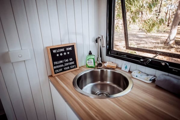 Una cocina compacta con un lavamanos de acero inoxidable, encimera de madera, una pizarra con un mensaje de bienvenida y vistas a árboles a través de una ventana.
