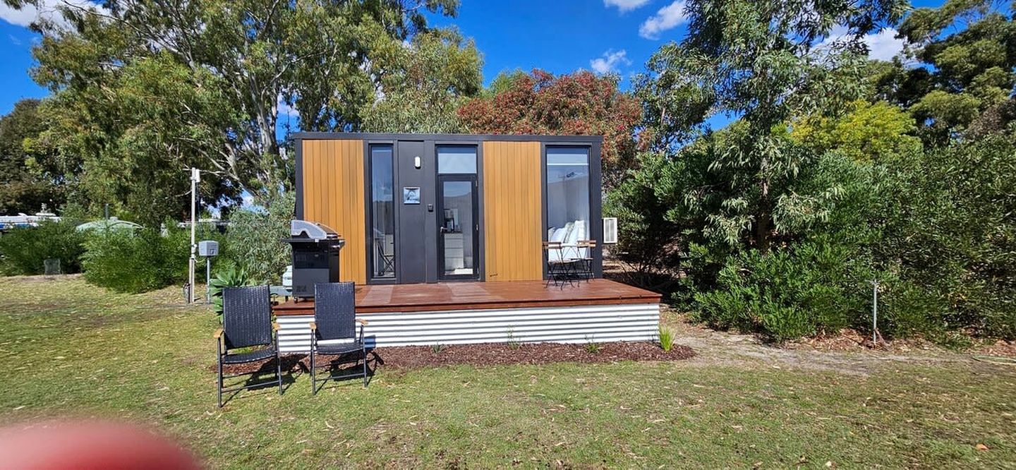 Un edificio moderno de una sola planta con techo plano, una terraza con dos sillas y una parrilla pequeña al aire libre.