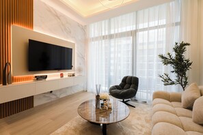 Ein modernes Wohnzimmer mit einem Flachbildfernseher, einem runden Couchtisch, einer Couch und einem großen Fenster mit transparenten Vorhängen.