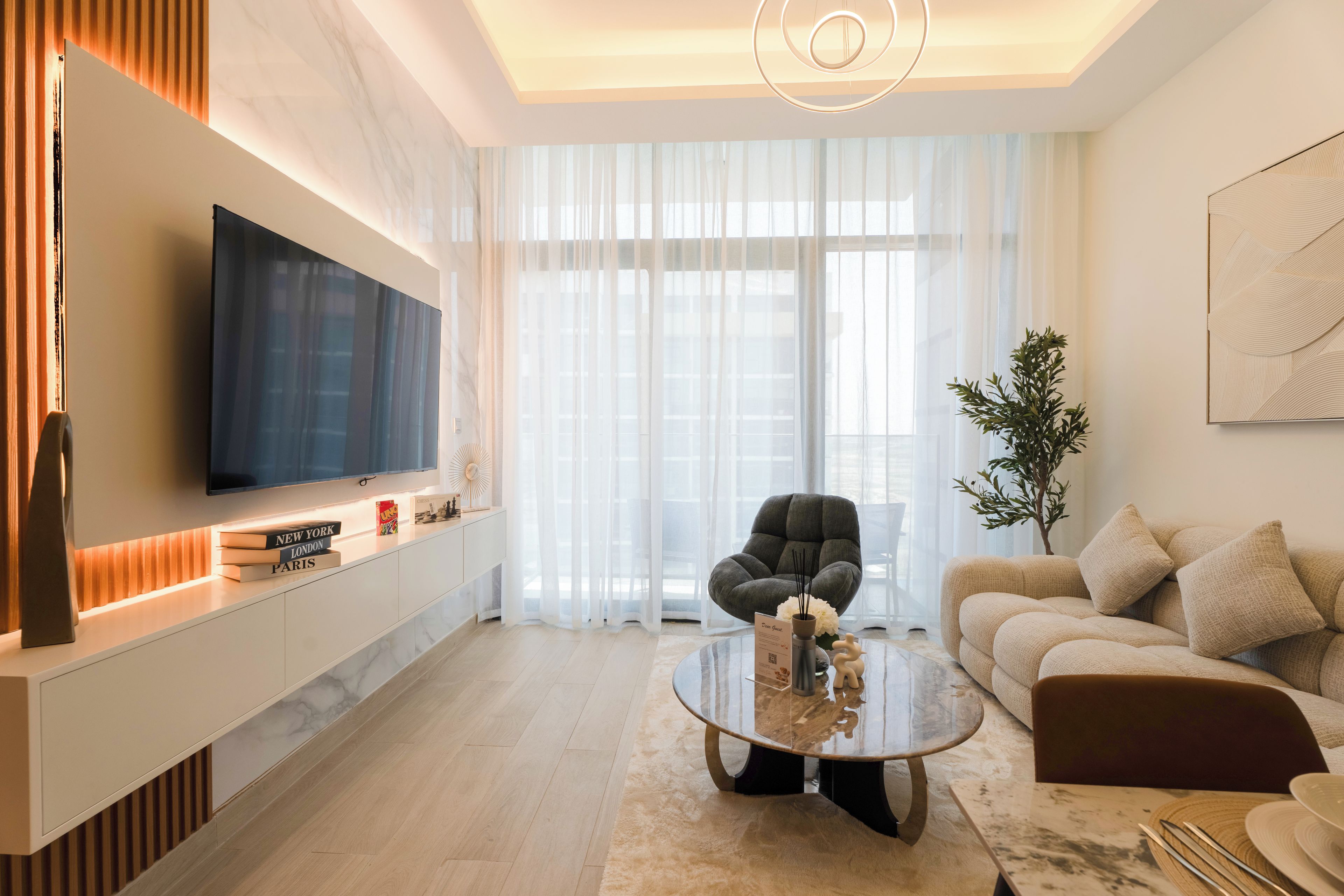 Een moderne woonkamer met een flatscreen televisie, een bank, een salontafel en een groot raam met dunne gordijnen.