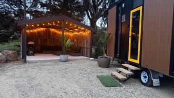 A wooden cabin with a covered porch and a yellow door, surrounded by trees and gravel.