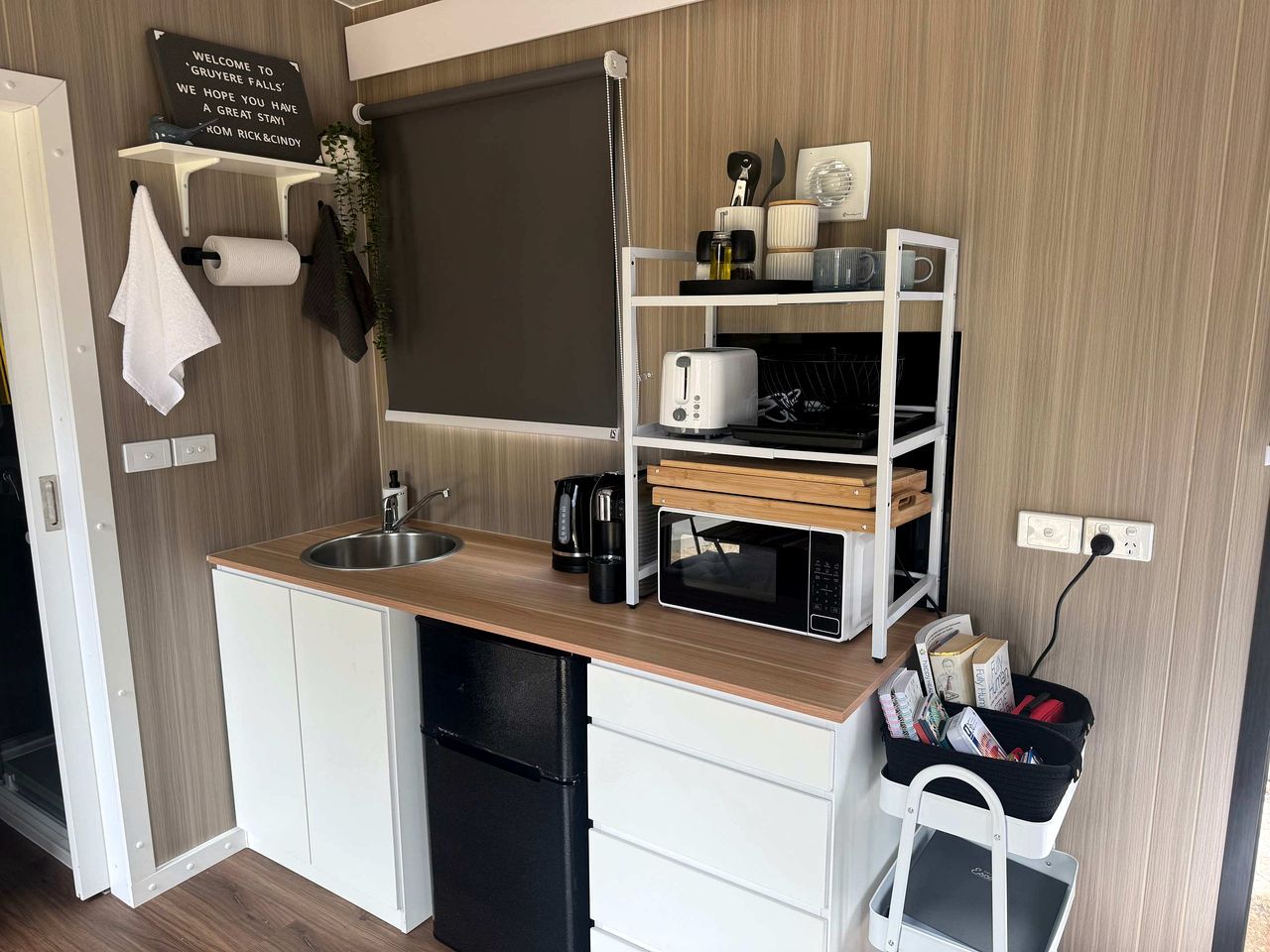 A compact kitchenette with a sink, a microwave, a coffee maker, and a small shelf with various items.