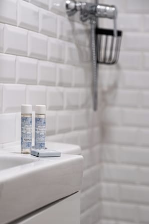 Une salle de bain avec des carreaux blancs, une pomme de douche et deux flacons de produits pour les soins des cheveux et du corps, posés sur un comptoir blanc.
