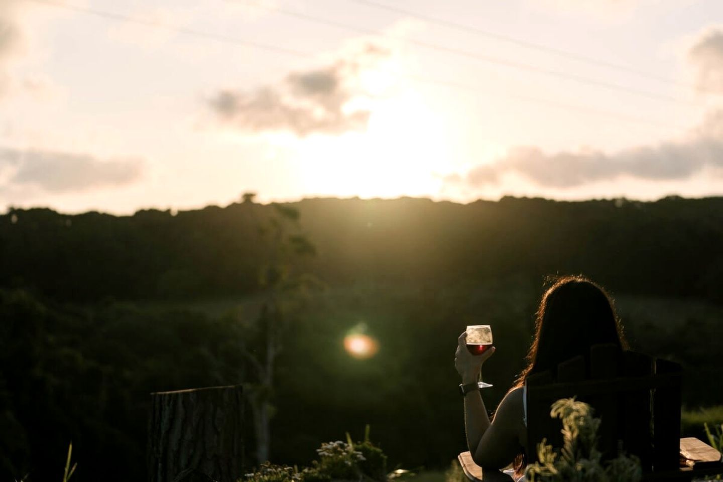 A person sitting outdoors with a cup, sunlight in the background, and vegetation around.
