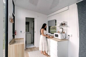 A compact kitchen with white cabinetry, a microwave, and a person preparing food. - Tiny House for Outdoor Adventure near Glacier Rock in Cairns, Queensland (Kuranda)