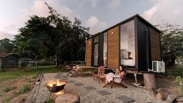 Een moderne blokhut met een vuurkuil, buitenmeubels en een groot raam met uitzicht op een natuurlandschap.