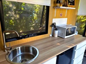 A compact kitchen with a stainless steel sink, a microwave, and a window with a view of greenery. - Sunshine Coast Glamping in a Tiny House for a Nature Escape in Queensland (Palmwoods)