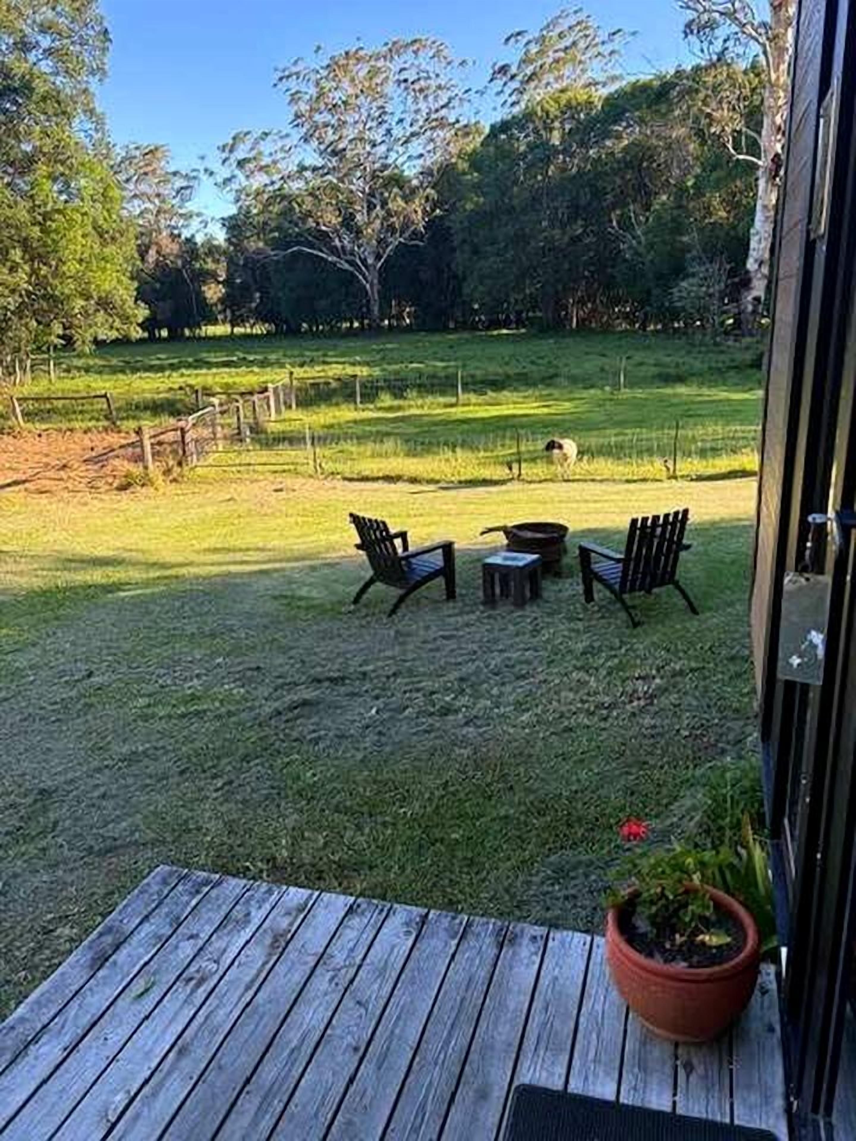 An outdoor patio with a wooden table and two chairs, surrounded by a grassy area and trees.