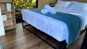 Een bed met een metalen frame, witte beddengoed en een blauwgroene plaid. Er is een houten plank met een wit object erop, en er is een plant zichtbaar door een raam.