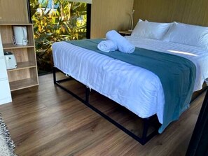 Una cama con estructura metálica, ropa de cama blanca y una manta color turquesa. Se observa un estante de madera con un objeto blanco y una planta visible a través de una ventana.