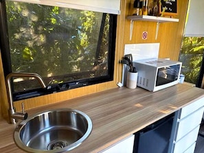 A compact kitchen with a stainless steel sink, a microwave, and a window with a view of greenery.