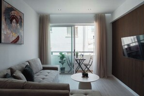A modern living room with a sofa, a coffee table, a TV mounted on the wall, and a balcony with a view. - Serene 2BR Mackenzie Apt 5 Min Walk to the Beach (Larnaka)