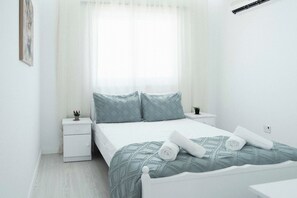 A minimalist bedroom with a bed, two nightstands, a window with sheer curtains, and a wall-mounted air conditioning unit. - Serene 2BR Mackenzie Apt 5 Min Walk to the Beach (Larnaka)