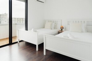 Une chambre moderne avec deux lits blancs, une table de chevet, une lampe et une grande fenêtre donnant sur des immeubles.