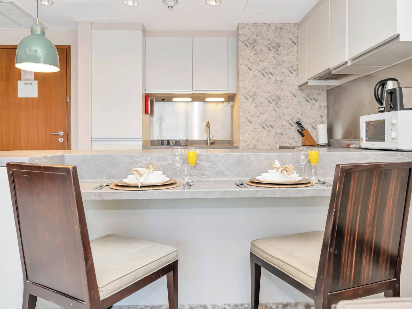 Une cuisine moderne avec un plan de travail en marbre, deux chaises en bois, une table à manger dressée pour deux personnes et un four à micro-ondes.