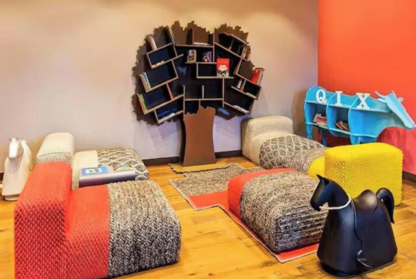 A room with a unique bookshelf designed to look like a tree, multiple cushioned seating areas, and a small blue play area.