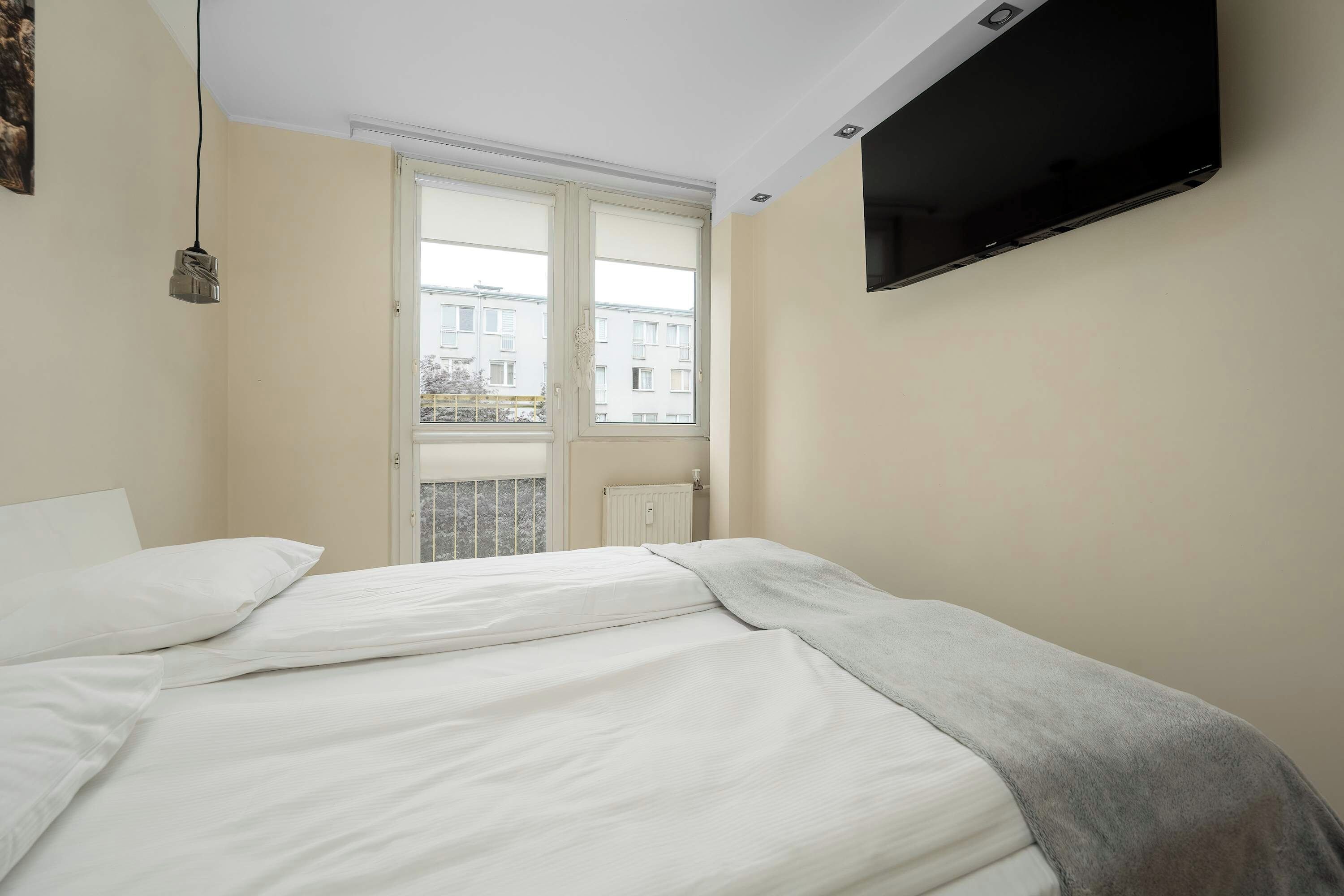 A bedroom with a bed, a television mounted on the wall, and a window with a view of buildings.
