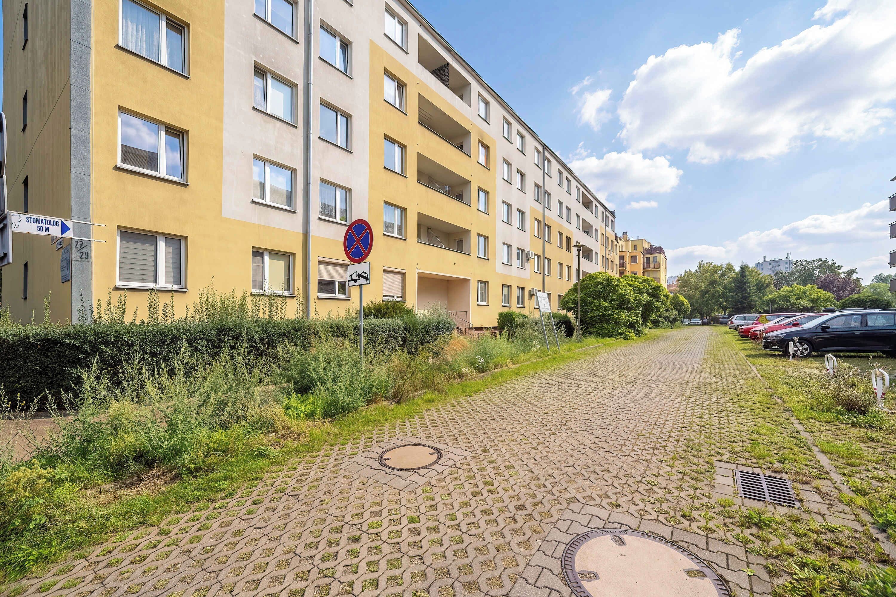 A paved pathway leading to a yellow multi-story residential building with a no parking sign, a manhole cover, and a row of parked cars.