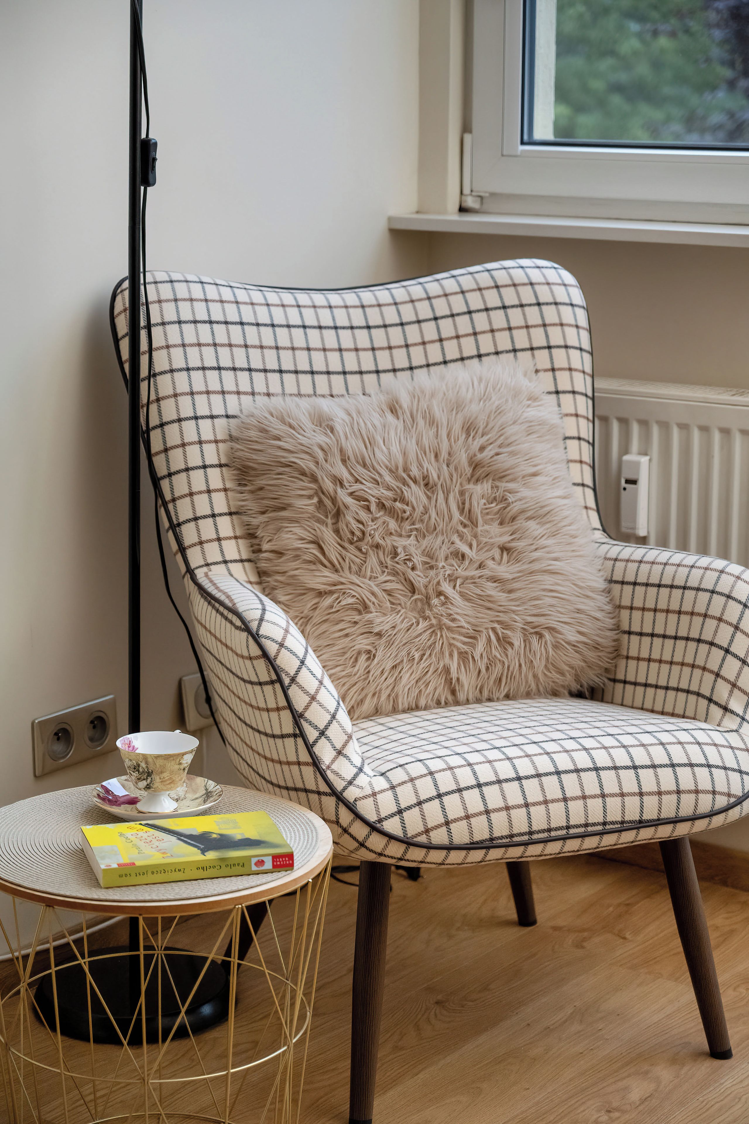 Tuoli, jossa on ruutukuviollinen kangas, ja pehmeä tyyny, pieni pyöreä pöytä, jonka päällä on kirja ja kulho, sekä taustalla näkyvä patteri.