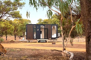 A modern, compact mobile home with a dark exterior, large windows, and a flat roof, situated in a dry, open landscape with sparse trees.