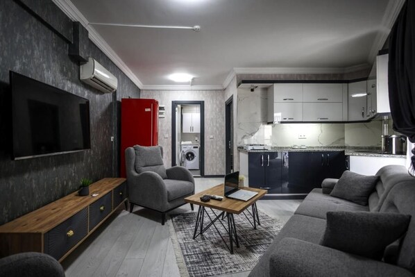 Una sala moderna con una televisión de pantalla plana, sofás grises, una mesa de centro y una cocina con armarios blancos y un electrodoméstico rojo.