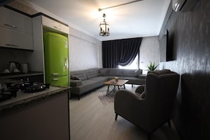 A modern living room with a sectional sofa, a coffee table, and a dining area with a green refrigerator. - Çınar Suit Hotel (Buca)