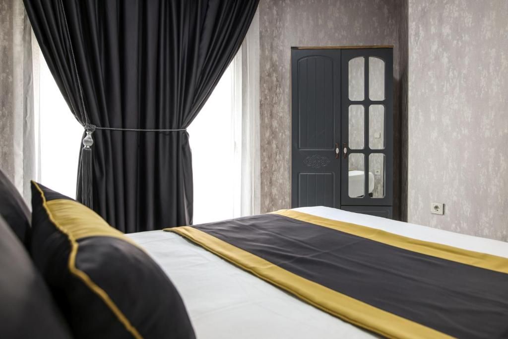 Quarto de hotel com cama, cortinas pretas, porta com painéis de vidro e parede texturizada.