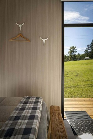 A bedroom with a bed, a wooden bench, a hanging coat rack, and a view of a grassy field through a large window.