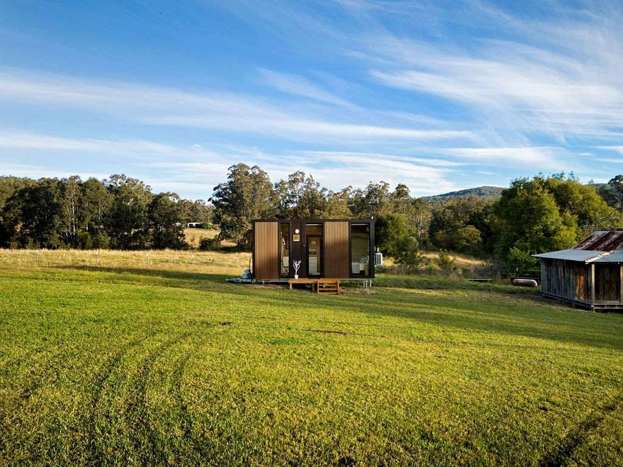 Une structure moderne de style cabane, dotée d’une terrasse en bois et d’un banc, entourée d’un champ herbeux et d’arbres.