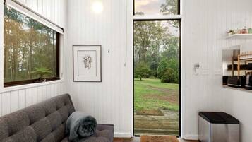 Quarto com sofá cinzento, um quadro emoldurado, tapete pequeno, porta que dá para um jardim e uma lixeira.